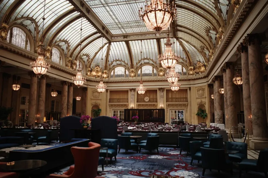A grand hotel dining room under a glass dome, with crystal chandeliers, marble columns and empty tables.